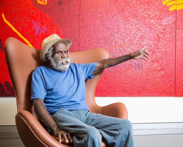 Artist Tommy Watson sitting in a tan lounge chair in front of a red wall