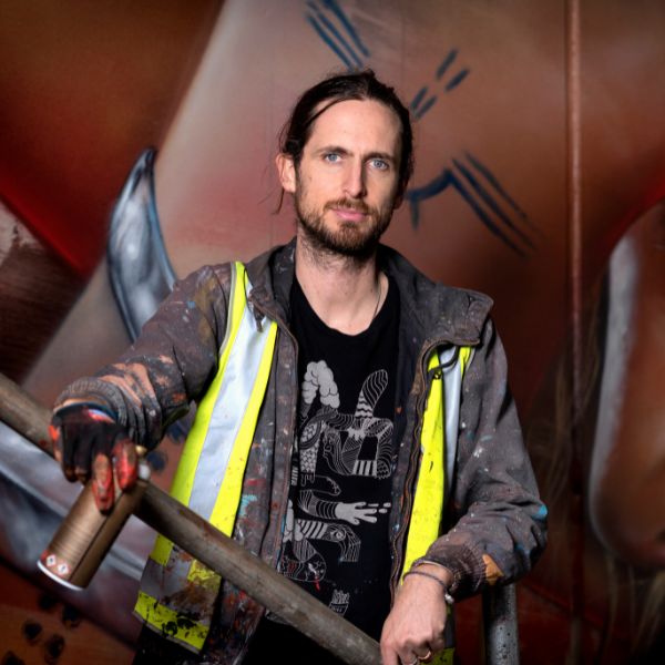 A dark-haired man holding spray paint and wearing a paint-stained shirt stands in front of an artwork