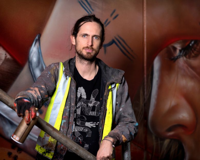 Matt Adnate, an artist with dark hair and paint splatters on his clothes, stands in front of a mural 