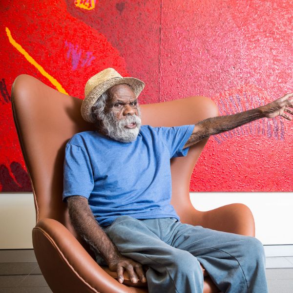 A man wearing a beige hat and blue shirt sits in front of a red mural