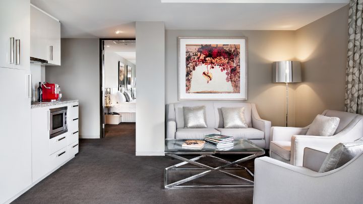 A grey and white hotel lounge room with a small kitchenette and three sofa chairs